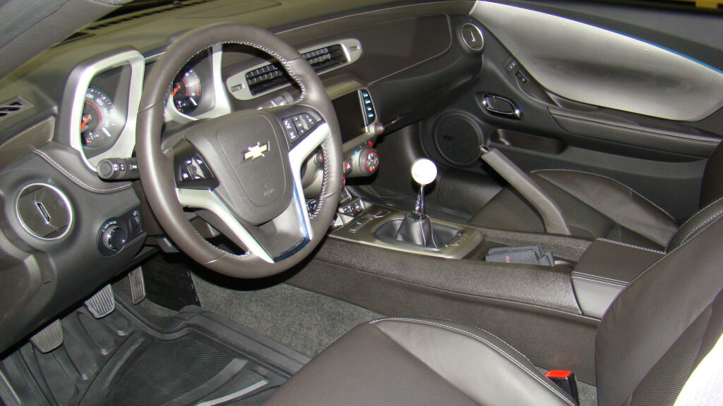 Interior of a Chevrolet Camaro showing black leather steering wheel with silver Chevrolet logo, dual analog gauges, dark multimedia screen, three round climate dials, and manual gear shift with white knob and leather boot.