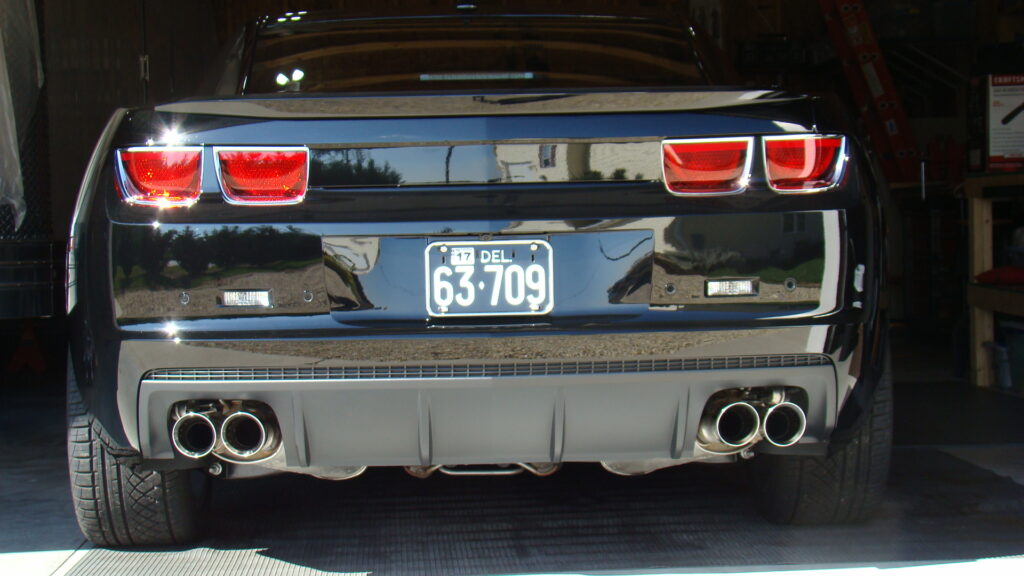 Rear view of a black Chevrolet Camaro with glossy paint, red tail lights, dual exhausts on each side, and a Delaware license plate, parked inside a garage with shelves, tools, and a red ladder visible.