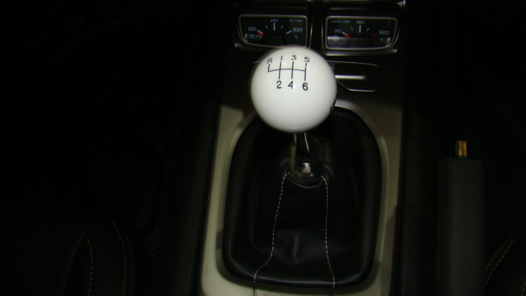Close-up of a car’s center console showing a white 6-speed manual gear shift knob with black pattern markings, black leather boot with white stitching, and two small round gauges with chrome bezels above.