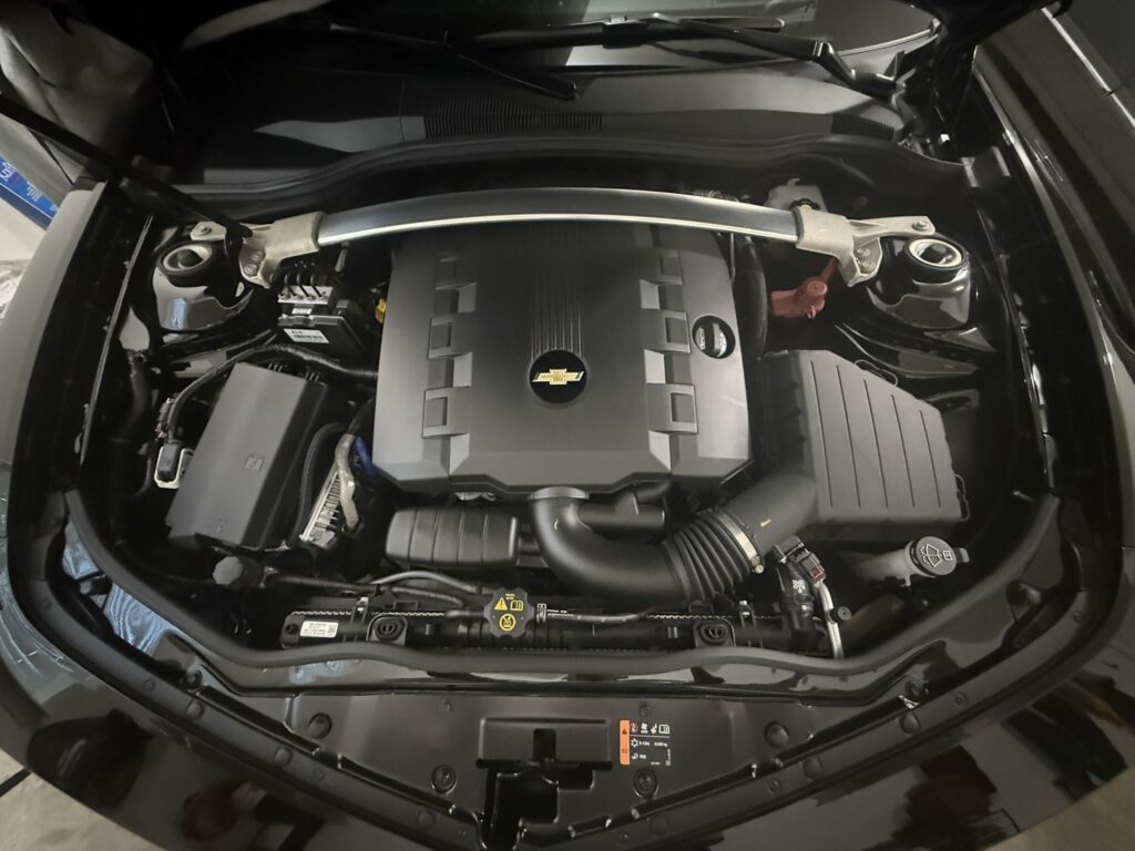 Chevrolet engine bay with black plastic engine cover featuring gold Chevrolet logo, silver strut bar across top, visible battery, coolant reservoir, and air intake components.